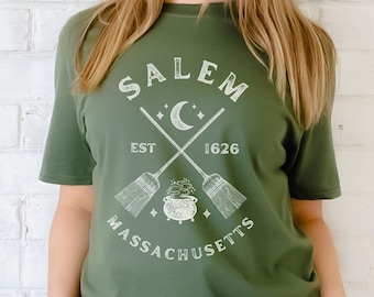 Puede incluir: Una camiseta verde con un diseño gráfico blanco que presenta el texto "Salem, Est. 1626, Massachusetts" con una luna creciente, escobas cruzadas y un caldero.