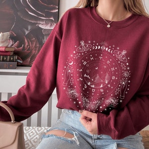 May include: A maroon sweatshirt with a white graphic design featuring a circle with celestial symbols, including moons, stars, zodiac signs, butterflies, and hands reaching towards the sky.
