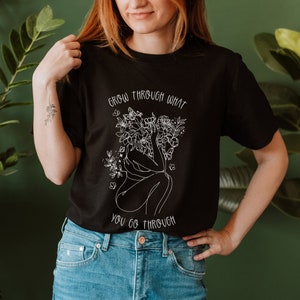 May include: Black t-shirt with a white graphic of a woman with flowers growing from her head. The text "Grow through what you go through" is printed on the shirt.