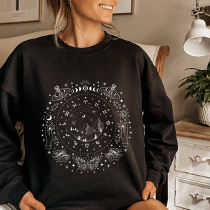 May include: Black sweatshirt with a white graphic design featuring a circle of celestial symbols, including moons, stars, butterflies, and zodiac signs. The design also includes a mountain range and two hands reaching towards the sky.