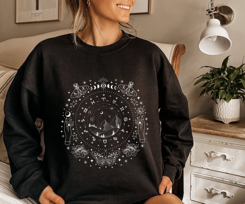 May include: Black sweatshirt with a white graphic design featuring a circle of celestial symbols, including moons, stars, butterflies, and zodiac signs. The design also includes a mountain range and two hands reaching towards the sky.