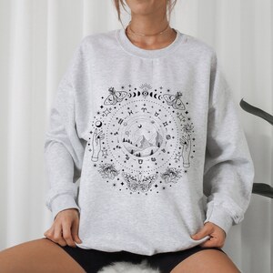 May include: Gray sweatshirt with a black and white graphic print of a celestial scene with mountains, stars, moons, butterflies, and hands. The graphic includes zodiac symbols.