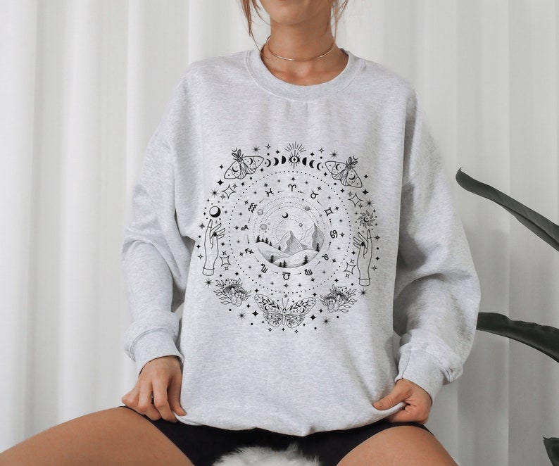 May include: Gray sweatshirt with a black and white graphic print of a celestial scene with mountains, stars, moons, butterflies, and hands. The graphic includes zodiac symbols.