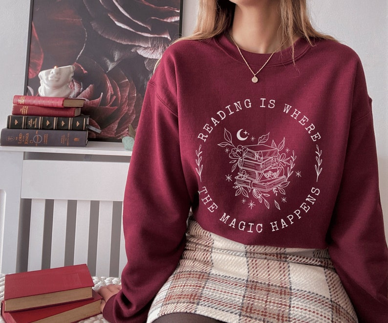 May include: Burgundy sweatshirt with a white graphic design that says "Reading is where the magic happens" with a crescent moon and a stack of books surrounded by flowers.