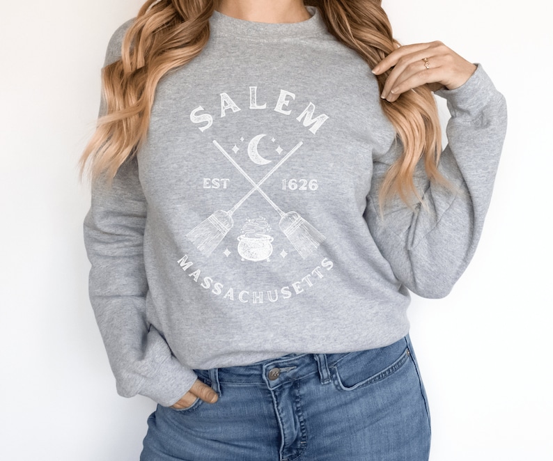 May include: Gray sweatshirt with a white graphic design that says "Salem, Est 1626, Massachusetts" with a crescent moon, two crossed brooms, and a cauldron.
