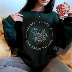 May include: A dark green sweatshirt with a white graphic design that says "Reading is where the magic happens" with a crescent moon, stars, and a stack of books.