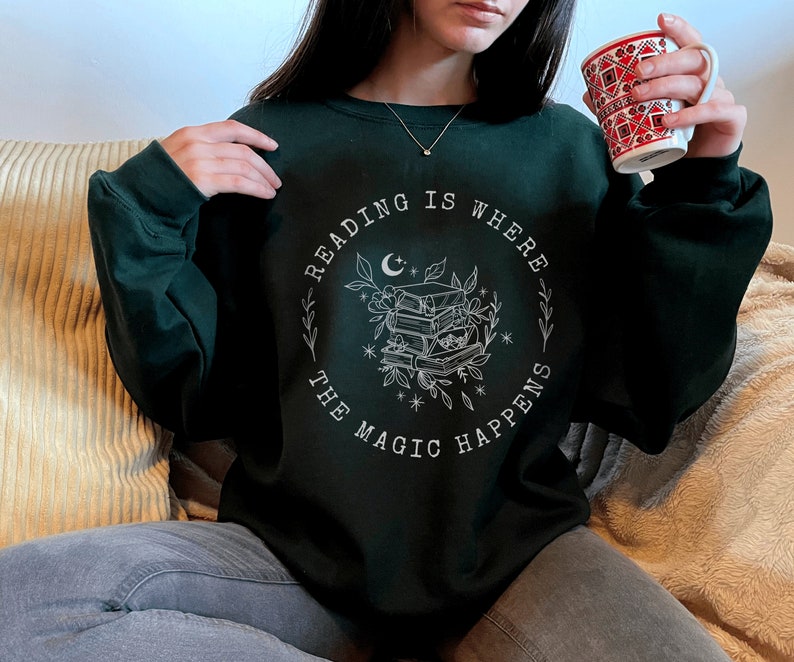 May include: A dark green sweatshirt with a white graphic design that says "Reading is where the magic happens" with a crescent moon, stars, and a stack of books.