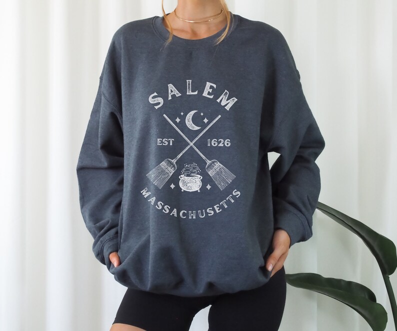 May include: A dark gray sweatshirt with a white graphic design featuring the text "SALEM EST 1626 MASSACHUSETTS" with a crescent moon, stars, and crossed broomsticks with a cauldron in the center.