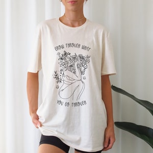 May include: A white t-shirt with a black line drawing of a person with flowers growing from their head. The text "Grow through what you go through" is printed on the shirt.
