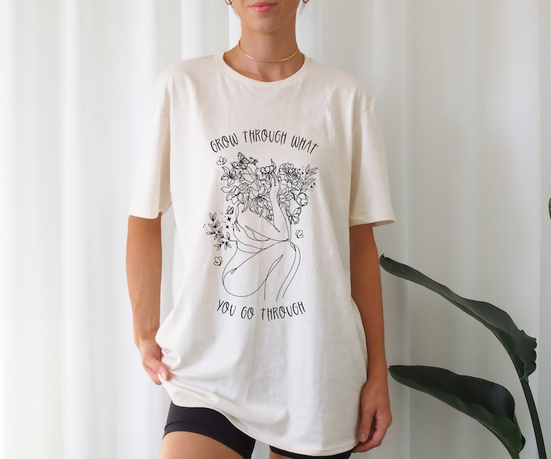 May include: A white t-shirt with a black line drawing of a person with flowers growing from their head. The text "Grow through what you go through" is printed on the shirt.