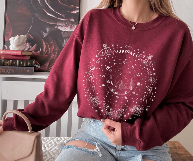 May include: A maroon sweatshirt with a white graphic design featuring a circle with celestial symbols, including moons, stars, zodiac signs, butterflies, and hands reaching towards the sky.