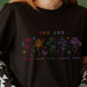 May include: Black t-shirt with a rainbow floral design and the text "YOU ARE" above the flowers. Below the flowers are the words "STRONG, BRAVE, LOVED, CAPABLE, SMART" in rainbow colors.