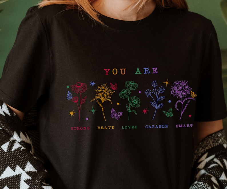 May include: Black t-shirt with a rainbow floral design and the text "YOU ARE" above the flowers. Below the flowers are the words "STRONG, BRAVE, LOVED, CAPABLE, SMART" in rainbow colors.