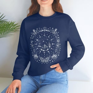 May include: Navy blue long-sleeved sweatshirt with a white graphic design of a circle with celestial symbols, including moons, stars, butterflies, and zodiac signs. The design also includes a mountain range and two hands reaching up.