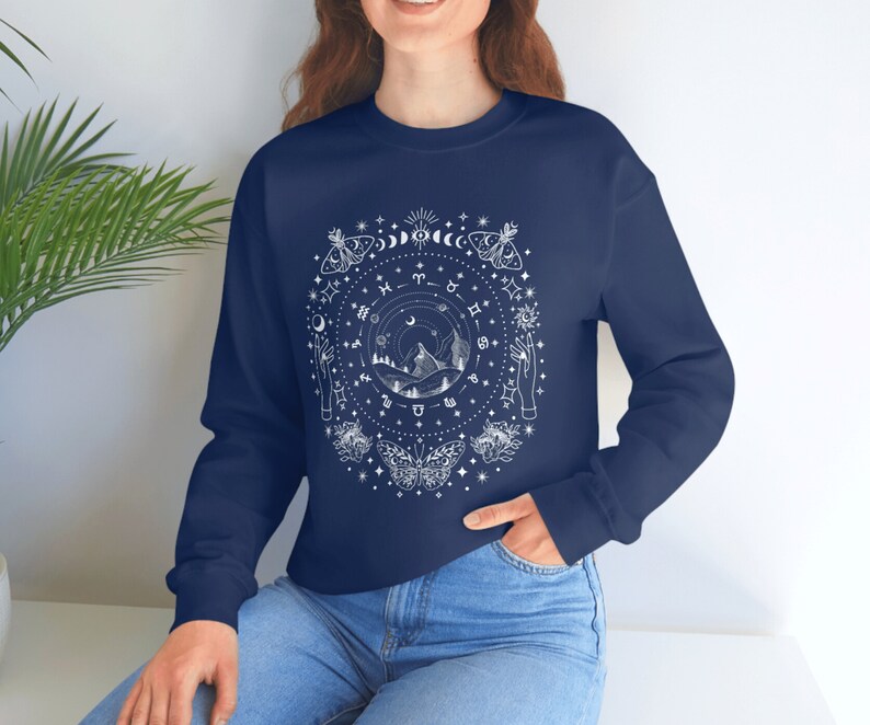 May include: Navy blue long-sleeved sweatshirt with a white graphic design of a circle with celestial symbols, including moons, stars, butterflies, and zodiac signs. The design also includes a mountain range and two hands reaching up.