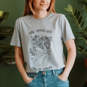 May include: A gray t-shirt with a black line drawing of a woman with flowers growing from her head. The text "Grow through what you go through" is printed on the shirt.