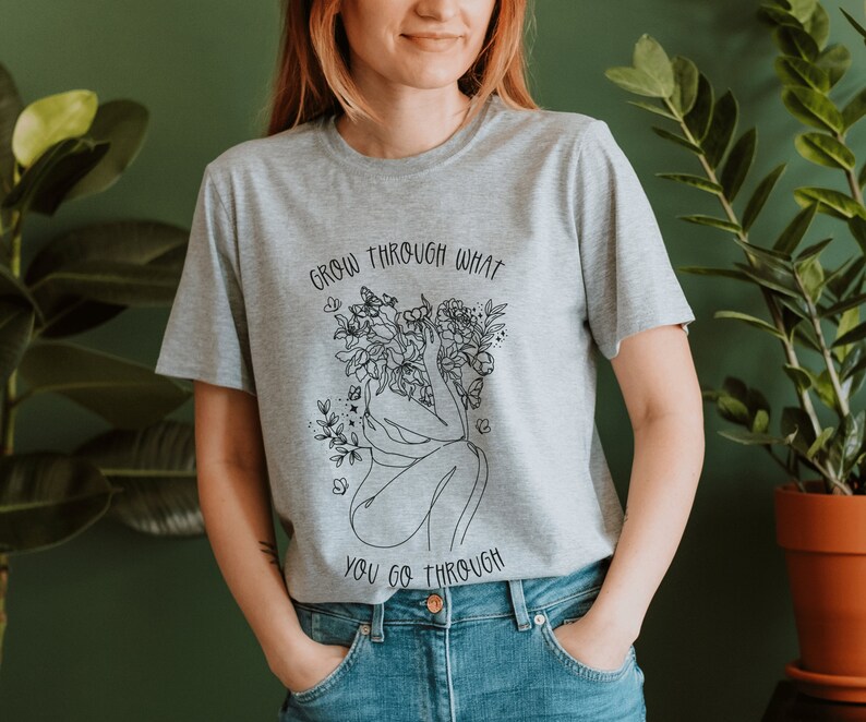 May include: A gray t-shirt with a black line drawing of a woman with flowers growing from her head. The text "Grow through what you go through" is printed on the shirt.