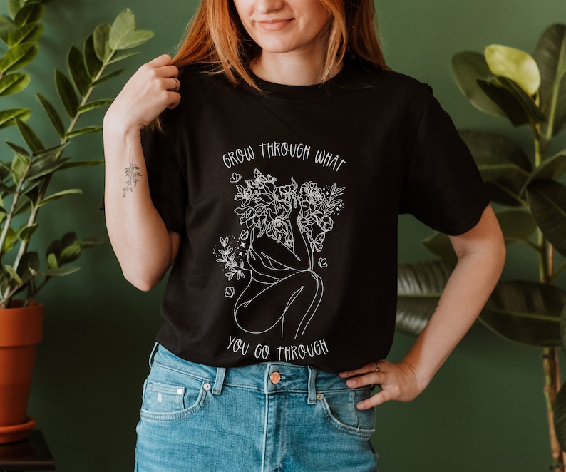 May include: Black t-shirt with a white graphic of a woman with flowers growing from her head. The text "Grow through what you go through" is printed on the shirt.