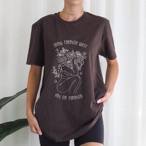May include: A brown t-shirt with a white graphic of a person surrounded by flowers and the text "grow through what you go through".