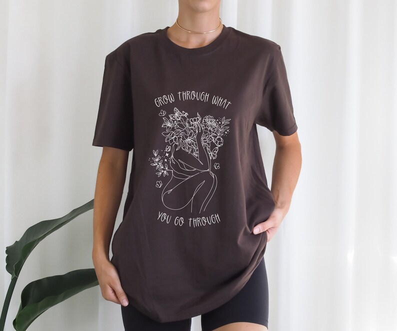 May include: A brown t-shirt with a white graphic of a person surrounded by flowers and the text "grow through what you go through".