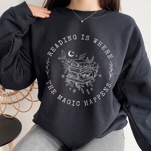 May include: Black sweatshirt with a white graphic design that says "Reading is where the magic happens" with a crescent moon and a stack of books surrounded by flowers.