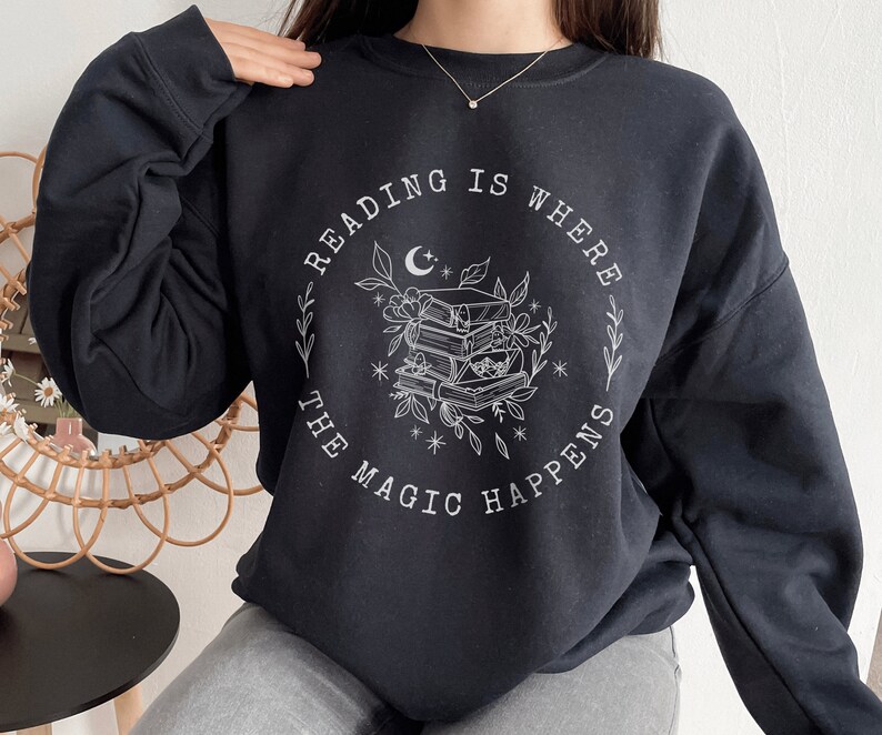 May include: Black sweatshirt with a white graphic design that says "Reading is where the magic happens" with a crescent moon and a stack of books surrounded by flowers.