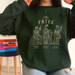 May include: A dark green sweatshirt with a graphic print of three figures representing the Fates, Clotho, Lachesis, and Atropos. The text "The Fates" is printed above the figures.