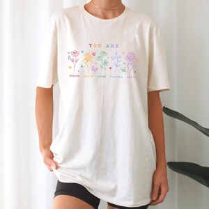 May include: A white t-shirt with a graphic design of flowers and butterflies. The text "YOU ARE" is printed above the flowers. Below the flowers are the words "STRONG, BRAVE, LOVED, CAPABLE, SMART".