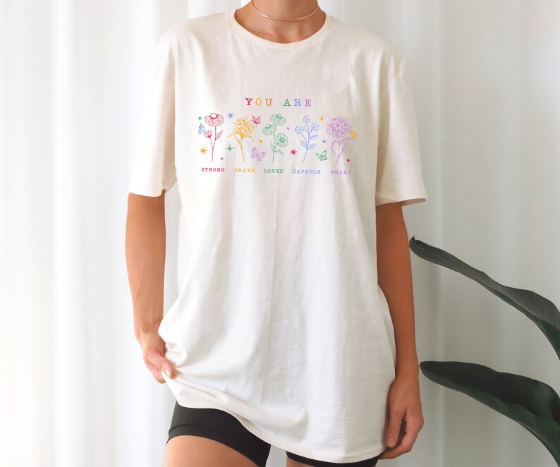 May include: A white t-shirt with a graphic design of flowers and butterflies. The text "YOU ARE" is printed above the flowers. Below the flowers are the words "STRONG, BRAVE, LOVED, CAPABLE, SMART".