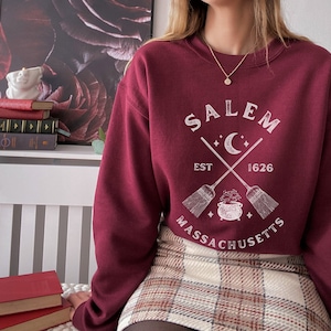 May include: A maroon sweatshirt with a white graphic design that says "Salem, Est 1626, Massachusetts" with a crescent moon and two crossed broomsticks with a cauldron in the center.
