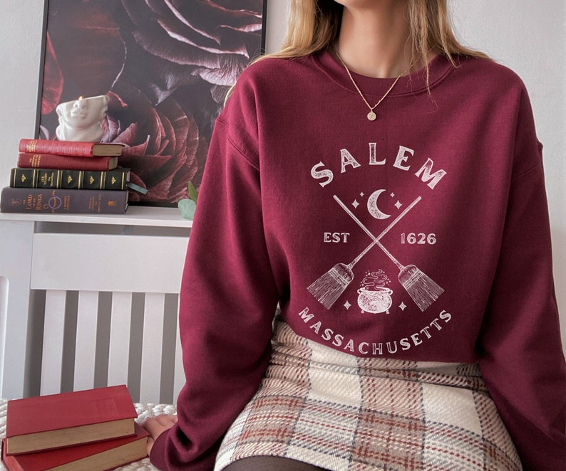 May include: A maroon sweatshirt with a white graphic design that says "Salem, Est 1626, Massachusetts" with a crescent moon and two crossed broomsticks with a cauldron in the center.