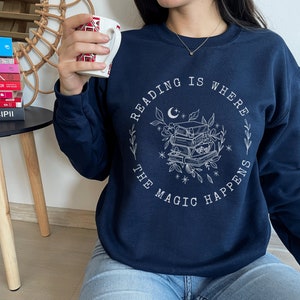 May include: A navy blue sweatshirt with a white graphic design that says "Reading is where the magic happens". The graphic features a stack of books, a crescent moon, and stars.
