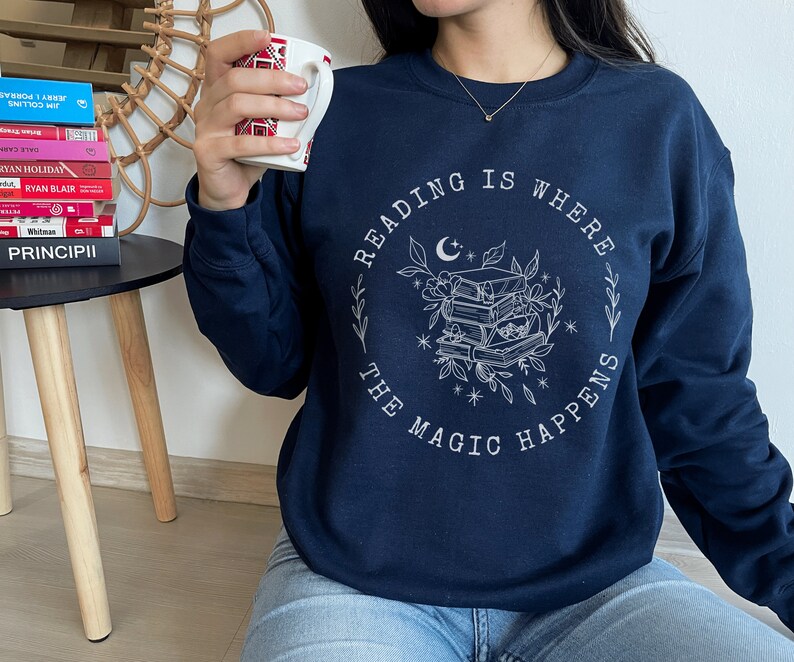 May include: A navy blue sweatshirt with a white graphic design that says "Reading is where the magic happens". The graphic features a stack of books, a crescent moon, and stars.