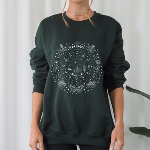 May include: A dark green sweatshirt with a white graphic print of a circle with celestial symbols, mountains, and moths. The graphic includes the zodiac signs, moons, stars, and hands.