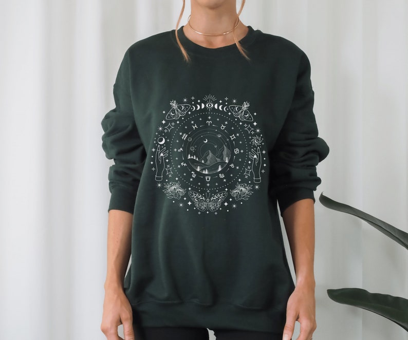 May include: A dark green sweatshirt with a white graphic print of a circle with celestial symbols, mountains, and moths. The graphic includes the zodiac signs, moons, stars, and hands.