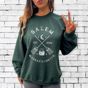May include: A dark green sweatshirt with a white graphic design featuring the text "Salem Massachusetts Est 1626" with a crescent moon, stars, and crossed broomsticks with a cauldron in the center.