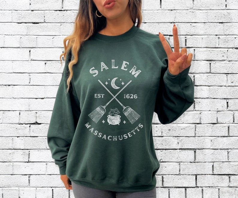 May include: A dark green sweatshirt with a white graphic design featuring the text "Salem Massachusetts Est 1626" with a crescent moon, stars, and crossed broomsticks with a cauldron in the center.