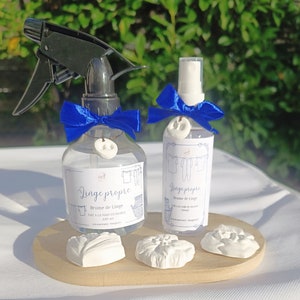 May include: Two clear glass spray bottles with blue ribbon bows and white ceramic charms. The bottles are labeled "Linge propre" and contain a linen spray. The bottles are on a wooden surface with white ceramic flowers.
