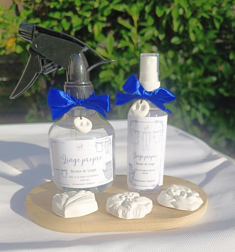 May include: Two clear glass spray bottles with blue ribbon bows and white ceramic charms. The bottles are labeled "Linge propre" and contain a linen spray. The bottles are on a wooden surface with white ceramic flowers.