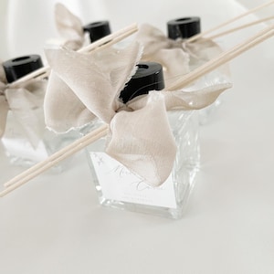 Könnte beinhalten: Drei durchsichtige Glasflaschen mit schwarzen Deckeln und Holzstäbchen. Jede Flasche ist mit einem beigefarbenen Band gebunden und hat ein Etikett mit der Aufschrift "Merci" und einem floralen Design.