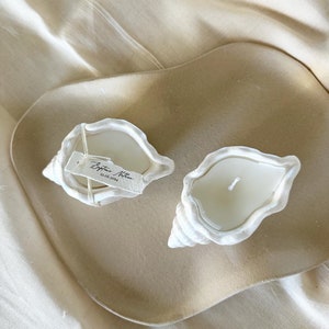 Könnte beinhalten: Zwei weiße Kerzen in Muschelform auf einem beigefarbenen Tablett. Die Kerzen sind unparfümiert und haben ein schlichtes Design. Das Tablett ist aus einem natürlichen Material gefertigt. Der Text auf dem Etikett lautet "Byttne Nathan 22.06.2024".