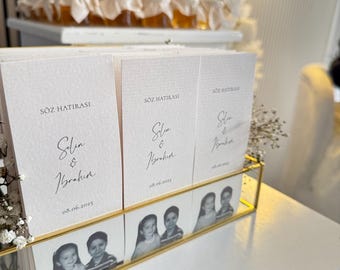 Puede incluir: Exhibición de recuerdos de boda con pequeños tarros de miel y tarjetas personalizadas. Las tarjetas, en un marco dorado, tienen el texto "SÖZ HATIRASI" y nombres con una fecha. El fondo incluye flores blancas y un letrero de neón.