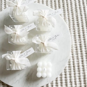 Op de afbeelding: Vijf kleine witte kaarsen in doorzichtige doosjes met witte strikken en gepersonaliseerde kaartjes met de tekst "Silen & Buben 02.12.2023". Een grotere witte kaars met een bubbelstructuur bevindt zich in het midden van de afbeelding.