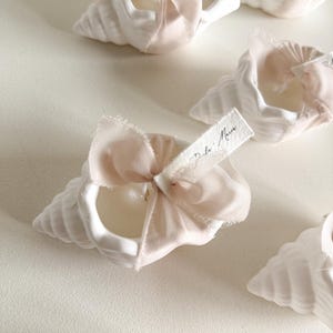 Könnte beinhalten: Weiße Muschelschalen-Kerzenhalter mit zarten rosa Schleifen und kleinen Etiketten. Die Kerzenhalter sind auf einer weißen Oberfläche angeordnet. Auf dem Etikett steht "Dala Maria".