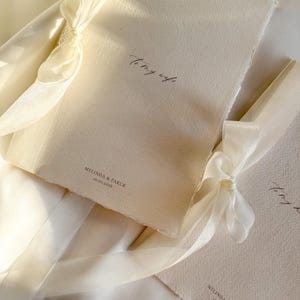 Op de afbeelding: Twee crèmekleurige papieren kaarten met geschulpte randen, vastgebonden met ivoorkleurige linten. De bovenste kaart leest "To my wife" in script. De onderste kaart heeft de namen "MELINDA & FARUK" en de datum "10.01.2026" afgedrukt.