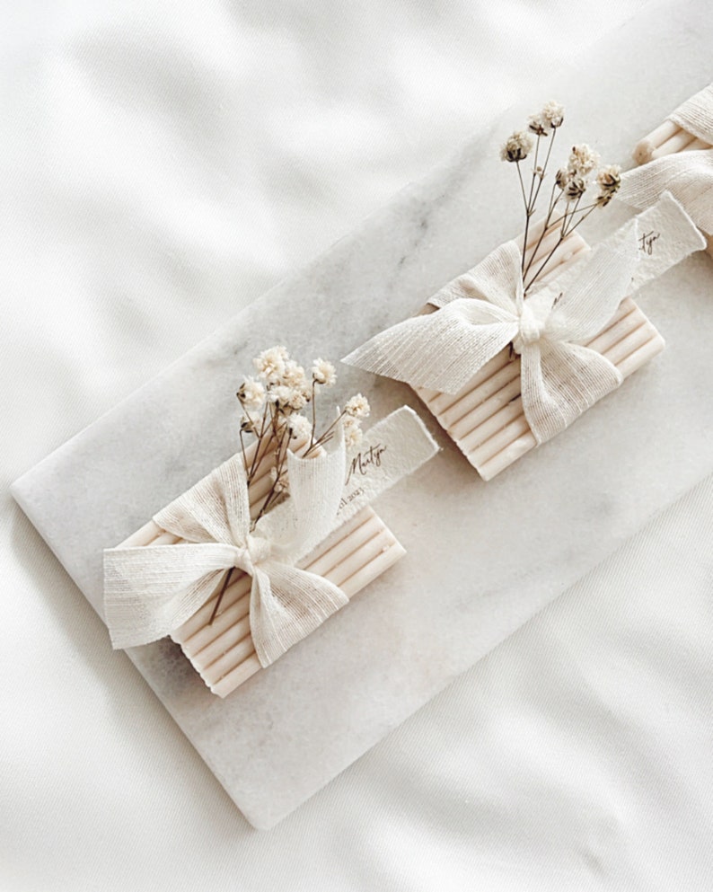 May include: Two small white bars of soap wrapped in white fabric with a small bouquet of dried white flowers and a handwritten tag that says "Misty" on top of a white marble surface.