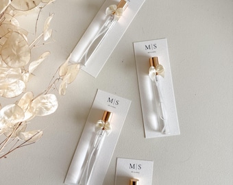 Op de afbeelding: Vier heldere glazen parfumverstuivers met goudkleurige doppen en kleine witte strikjes, elk bevestigd aan een witte kaart. De kaarten hebben de initialen "M/S" en de datum erop gedrukt. De verstuivers zijn op een lichtgekleurd oppervlak geplaatst.