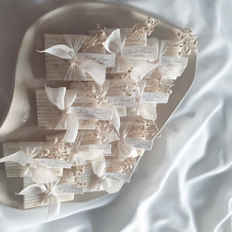 May include: A collection of small white soap favors tied with white ribbon and decorated with dried white flowers. Each favor has a small tag with the text "Jane & James" and the date "2023.05.14".