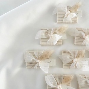 May include: A collection of small, rectangular boxes wrapped in white fabric with a bow and a sprig of dried grass. Each box has a small, handwritten tag attached.