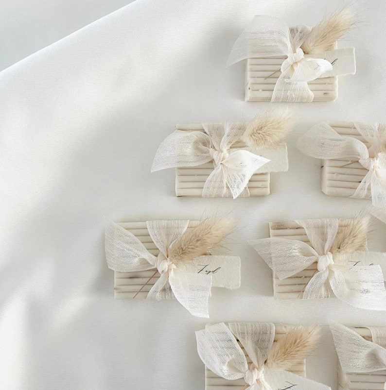 May include: A collection of small, rectangular boxes wrapped in white fabric with a bow and a sprig of dried grass. Each box has a small, handwritten tag attached.